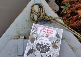 A graphic novel titled Idle Women on the water rests on front of a grey muddy canal boat